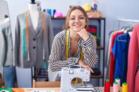 Téléchargez les photos : Jeune femme blonde tailleur souriant confiant appuyé sur la machine à coudre au magasin de tailleur - en image libre de droit