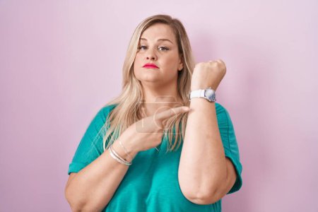 Photo for Caucasian plus size woman standing over pink background in hurry pointing to watch time, impatience, looking at the camera with relaxed expression - Royalty Free Image