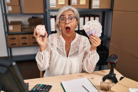 Photo for Middle age woman with grey hair working at small business ecommerce holding piggy bank and zloty afraid and shocked with surprise and amazed expression, fear and excited face. - Royalty Free Image