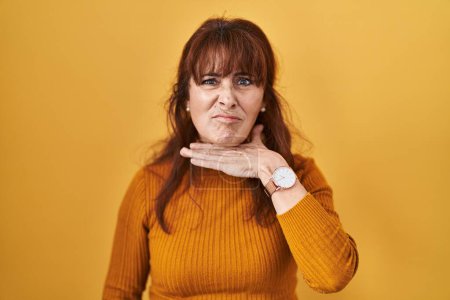 Téléchargez les photos : Femme hispanique du Moyen Âge debout sur fond jaune couper la gorge avec la main comme couteau, menacer d'agression avec une violence furieuse - en image libre de droit