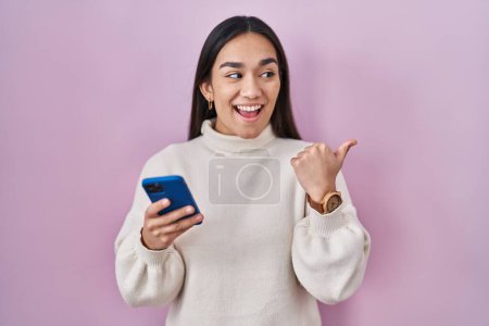 Photo for Young south asian woman using smartphone pointing thumb up to the side smiling happy with open mouth - Royalty Free Image