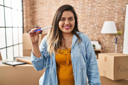 Photo for Young hispanic woman holding pregnancy test result at new home looking positive and happy standing and smiling with a confident smile showing teeth - Royalty Free Image