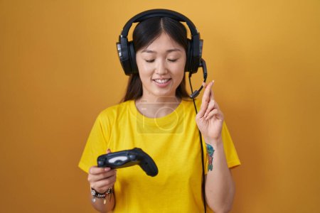Téléchargez les photos : Jeune femme chinoise jouant jeu vidéo tenant contrôleur geste doigt croisé souriant avec espoir et les yeux fermés. chance et concept superstitieux. - en image libre de droit