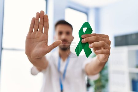 Foto de Young hispanic doctor man holding support green ribbon at the clinic with open hand doing stop sign with serious and confident expression, defense gesture - Imagen libre de derechos