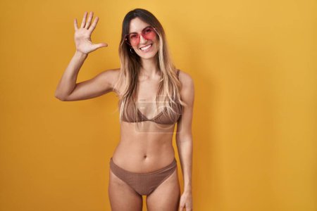 Téléchargez les photos : Jeune femme hispanique portant un bikini sur fond jaune montrant et pointant vers le haut avec les doigts numéro cinq tout en souriant confiant et heureux. - en image libre de droit