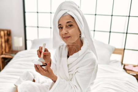 Foto de Mujer de pelo gris de mediana edad que aplica maquillaje en polvo sentado en la cama en el dormitorio - Imagen libre de derechos