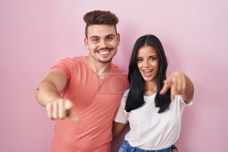 Foto de Joven pareja hispana de pie sobre fondo rosa señalándote a ti y a la cámara con los dedos, sonriendo positiva y alegre - Imagen libre de derechos