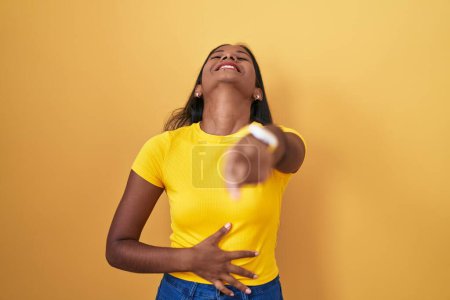 Foto de Young indian woman standing over yellow background laughing at you, pointing finger to the camera with hand over body, shame expression - Imagen libre de derechos