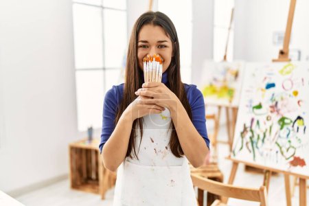 Photo for Young latin woman covering mouth with paintbrushes at art studio - Royalty Free Image