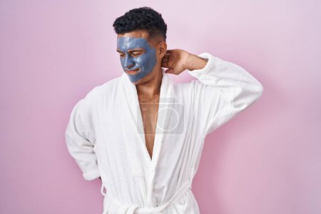 Téléchargez les photos : Jeune homme hispanique portant un masque de beauté et peignoir de bain souffrant d'une blessure au cou, toucher le cou avec la main, douleur musculaire - en image libre de droit
