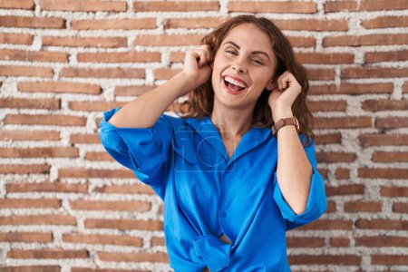 Photo for Beautiful brunette woman standing over bricks wall smiling pulling ears with fingers, funny gesture. audition problem - Royalty Free Image