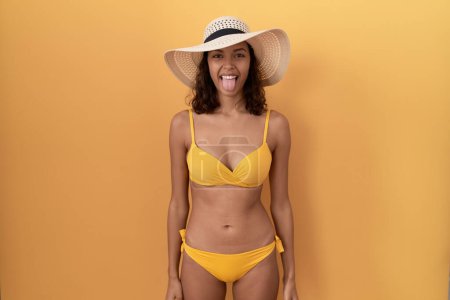 Foto de Mujer hispana joven con bikini y sombrero de verano sacando la lengua feliz con expresión divertida. concepto de emoción. - Imagen libre de derechos
