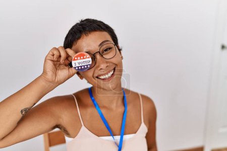 Foto de Joven mujer hispana sonriendo confiada sosteniendo insignia de voto trabajando en colegio electoral - Imagen libre de derechos