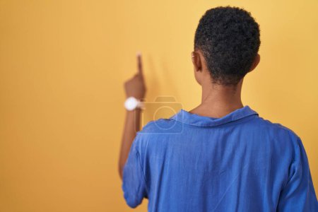 Téléchargez les photos : Femme afro-américaine debout sur fond jaune posant vers l'arrière pointant vers l'avant avec la main du doigt - en image libre de droit
