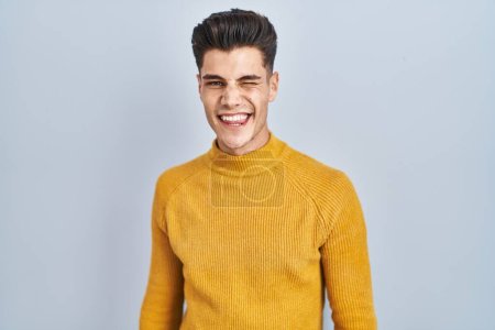 Téléchargez les photos : Jeune homme hispanique debout sur fond bleu clin d'oeil en regardant la caméra avec une expression sexy, gai et heureux visage. - en image libre de droit