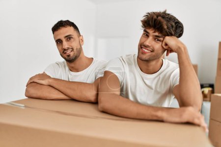 Foto de Dos hombres hispanos en pareja sonriendo confiados apoyados en una caja de cartón en un nuevo hogar - Imagen libre de derechos