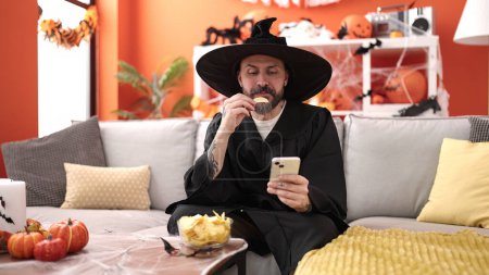 Foto de Joven hombre calvo usando disfraz de mago comiendo papas usando teléfono inteligente en casa - Imagen libre de derechos