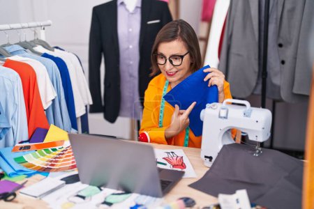 Téléchargez les photos : Femme d'âge moyen tailleur tenant tissu à l'aide d'un ordinateur portable au magasin de tailleur - en image libre de droit