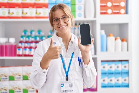 Téléchargez les photos : Jeune femme caucasienne travaillant à la pharmacie pharmacie montrant écran smartphone souriant heureux et positif, pouce levé faire excellent et signe d'approbation - en image libre de droit