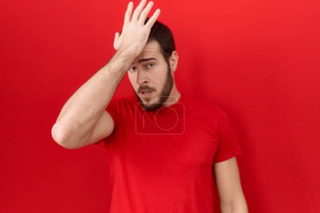 Foto de Joven hombre hispano con camiseta roja casual sorprendido con la mano en la cabeza por error, recordar el error. olvidado, mal concepto de memoria. - Imagen libre de derechos