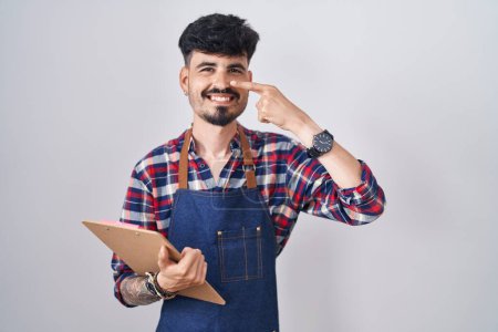 Téléchargez les photos : Jeune homme hispanique avec barbe portant tablier de serveur tenant presse-papiers pointant avec le doigt de la main vers le visage et le nez, souriant gai. concept de beauté - en image libre de droit