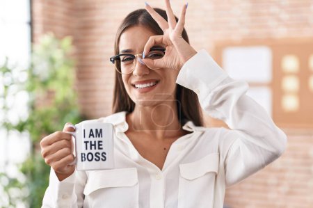 Photo for Young brunette woman drinking from i am the boss coffee cup smiling happy doing ok sign with hand on eye looking through fingers - Royalty Free Image
