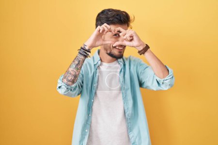 Photo for Young hispanic man with tattoos standing over yellow background doing heart shape with hand and fingers smiling looking through sign - Royalty Free Image