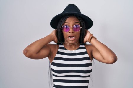 Téléchargez les photos : Jeune afro-américain avec des tresses portant chapeau et lunettes de soleil fou et effrayé avec les mains sur la tête, effrayé et surpris de choc avec la bouche ouverte - en image libre de droit