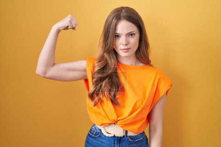 Caucasian woman standing over yellow background strong person showing arm muscle, confident and proud of power 