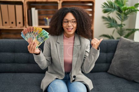 Foto de Joven mujer afroamericana sosteniendo dólares australianos en consulta oficina apuntando pulgar hacia el lado sonriendo feliz con la boca abierta - Imagen libre de derechos