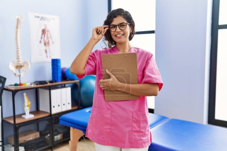 Téléchargez les photos : Jeune femme latine portant un uniforme de physiothérapeute tenant le presse-papiers à la clinique - en image libre de droit
