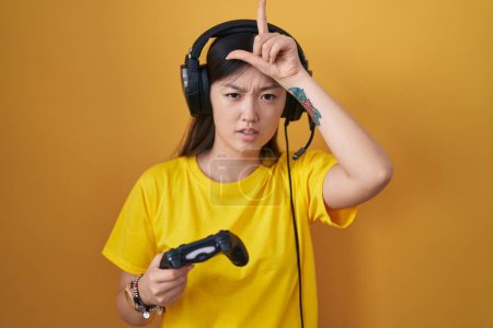 Photo for Chinese young woman playing video game holding controller making fun of people with fingers on forehead doing loser gesture mocking and insulting. - Royalty Free Image