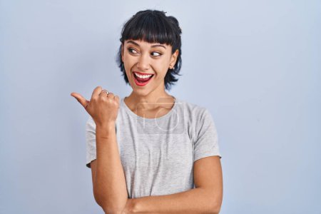 Photo for Young hispanic woman wearing casual t shirt over blue background smiling with happy face looking and pointing to the side with thumb up. - Royalty Free Image