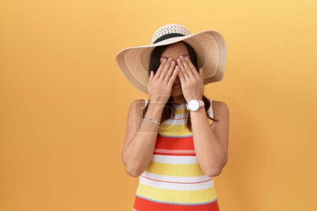 Foto de Mujer china de mediana edad con sombrero de verano sobre fondo amarillo frotando los ojos para la fatiga y el dolor de cabeza, la expresión somnolienta y cansada. problema de visión - Imagen libre de derechos