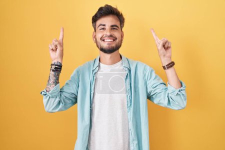 Foto de Young hispanic man with tattoos standing over yellow background smiling amazed and surprised and pointing up with fingers and raised arms. - Imagen libre de derechos