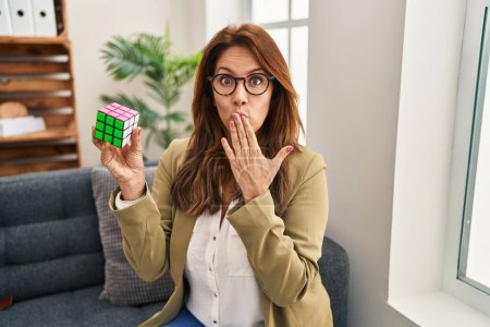 Foto de Hispanic woman playing colorful puzzle cube intelligence game covering mouth with hand, shocked and afraid for mistake. surprised expression - Imagen libre de derechos
