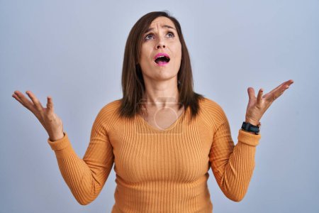 Foto de Middle age brunette woman standing wearing orange sweater crazy and mad shouting and yelling with aggressive expression and arms raised. frustration concept. - Imagen libre de derechos