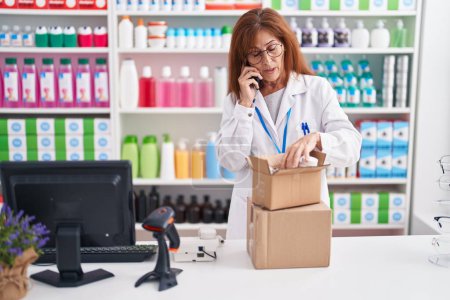 Téléchargez les photos : Pharmacien femme d'âge moyen parlant sur smartphone tenant bouteille de pilules à la pharmacie - en image libre de droit