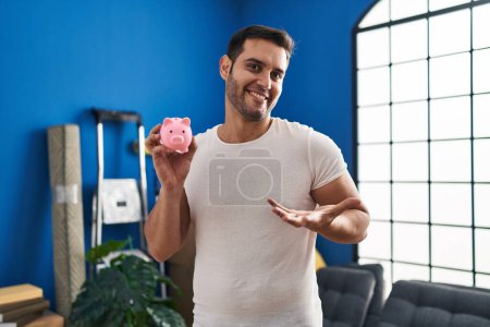 Téléchargez les photos : Jeune homme hispanique avec barbe tenant tirelire à la nouvelle maison célébrant la réalisation avec sourire heureux et expression gagnante avec la main levée - en image libre de droit