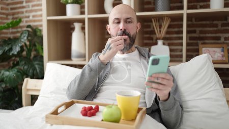 Foto de Joven hombre calvo desayunando usando smartphone en el dormitorio - Imagen libre de derechos