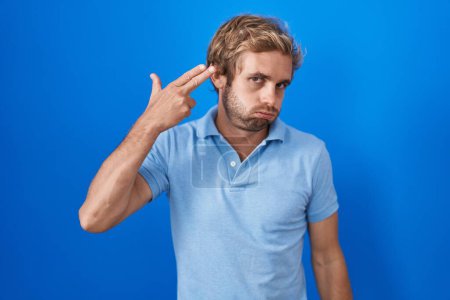 Photo for Caucasian man standing over blue background shooting and killing oneself pointing hand and fingers to head like gun, suicide gesture. - Royalty Free Image