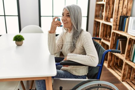 Photo for Middle age grey-haired woman using inhaler sitting on wheelchair at home - Royalty Free Image