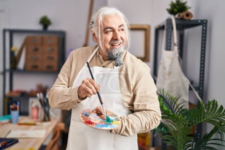 Photo for Middle age grey-haired man artist smiling confident holding paintbrush and palette at art studio - Royalty Free Image