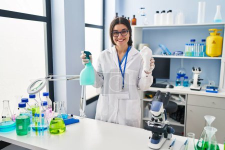 Téléchargez les photos : Young hispanic woman wearing scientist uniform cleaning using sanitizer gel at laboratory - en image libre de droit