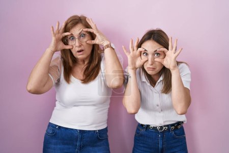 Foto de Madre e hija hispanas vistiendo una camiseta blanca casual sobre fondo rosa tratando de abrir los ojos con los dedos, soñolientas y cansadas por la fatiga matutina - Imagen libre de derechos
