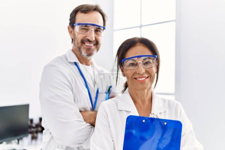 Photo for Middle age man and woman partners wearing scientist uniform holding clipboard with arms crossed gesture at laboratory - Royalty Free Image