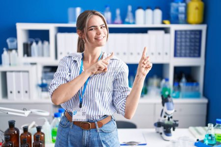 Foto de Joven mujer hermosa que trabaja en el laboratorio científico sonriendo y mirando a la cámara apuntando con dos manos y dedos hacia un lado. - Imagen libre de derechos