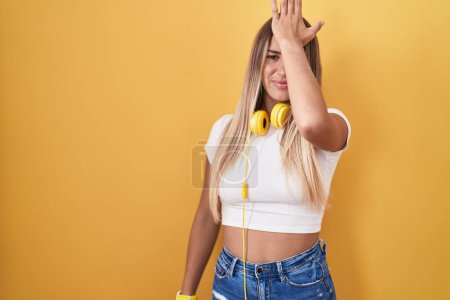 Téléchargez les photos : Jeune femme blonde debout sur fond jaune portant des écouteurs surpris avec la main sur la tête pour erreur, rappelez-vous erreur. oublié, mauvais concept de mémoire. - en image libre de droit