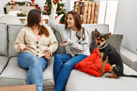 Foto de Dos mujeres conversando sentadas con el perro junto al árbol de navidad en casa - Imagen libre de derechos