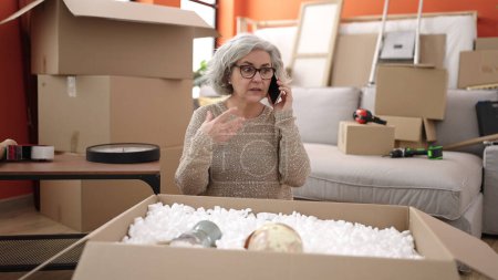Foto de Mujer de mediana edad con pelo gris hablando en el teléfono inteligente desembalaje caja de cartón en el nuevo hogar - Imagen libre de derechos